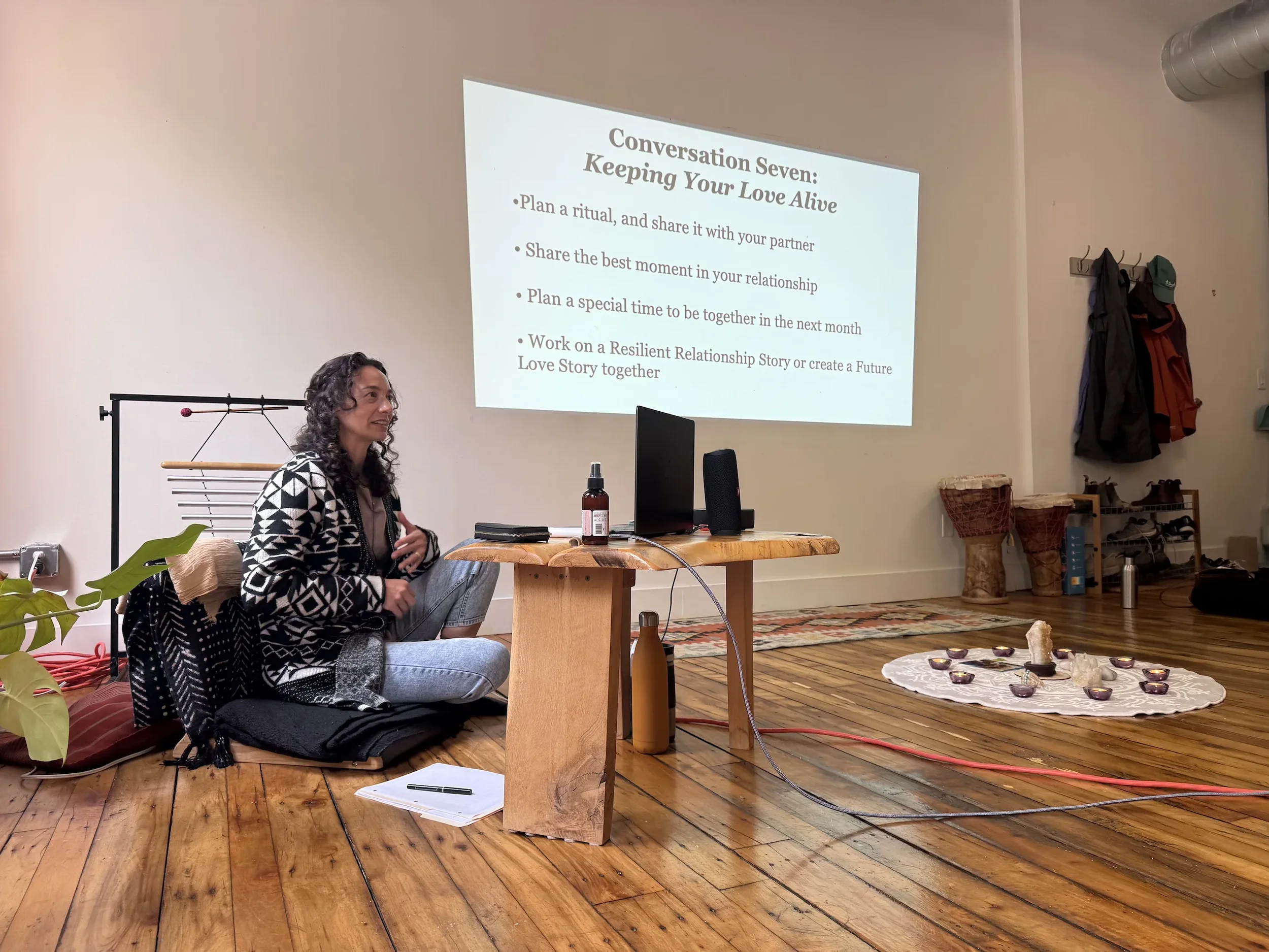 Alexis Lowenstein at a Hold Me Tight Workshop, sitting on the floor in front of a laptop and a projection screen displaying a presentation titled 'Conversation Seven: Keeping Your Love Alive,' listing five bullet points.