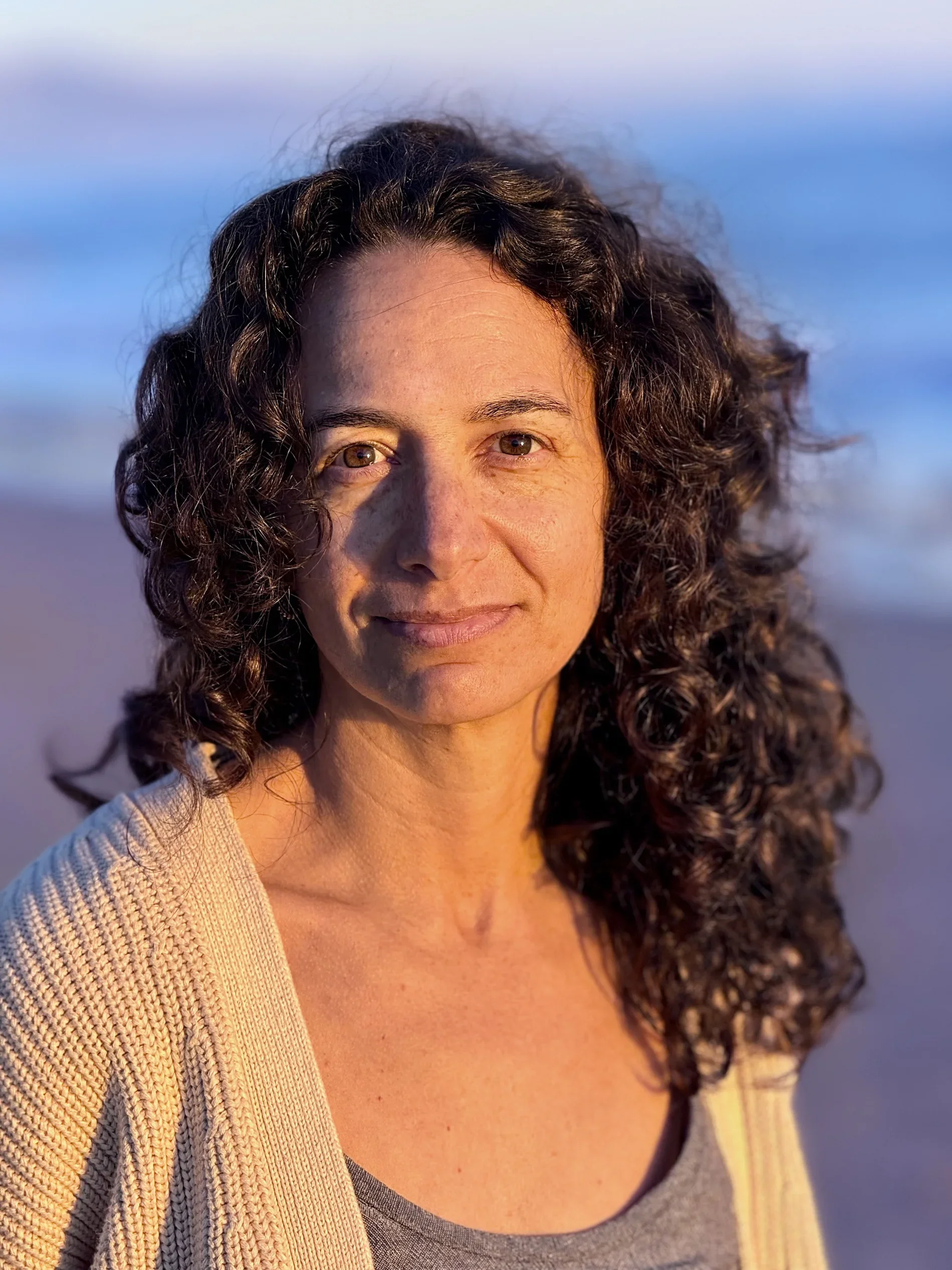 Close-up of Alexis Lowenstein outdoors with sunlight on her skin, curly hair, and wearing a gray top and a beige cardigan. Blurred ocean background.