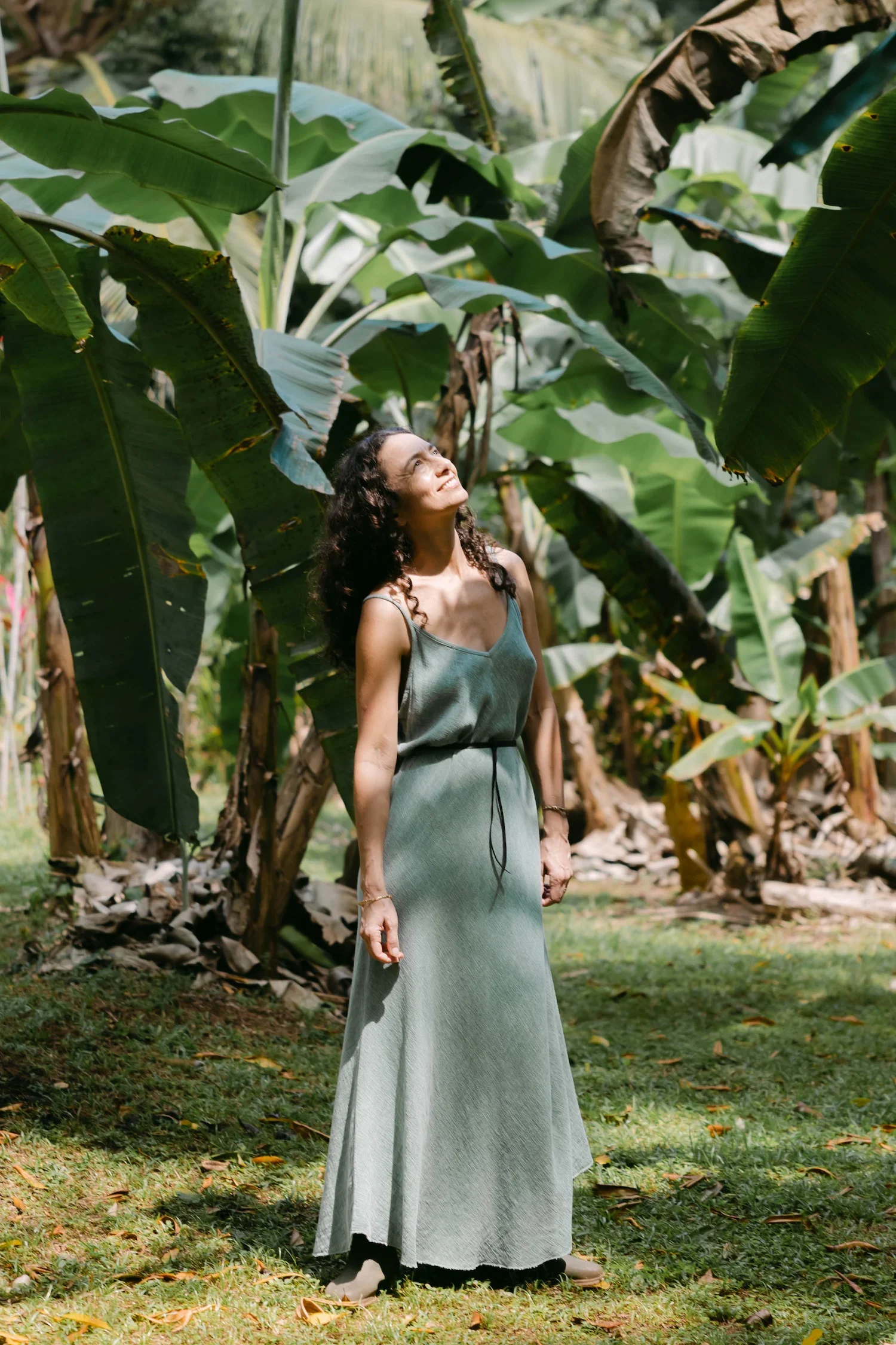 Alexis Lowenstein standing outdoors in a garden, surrounded by large banana leaves, looking upwards and smiling.