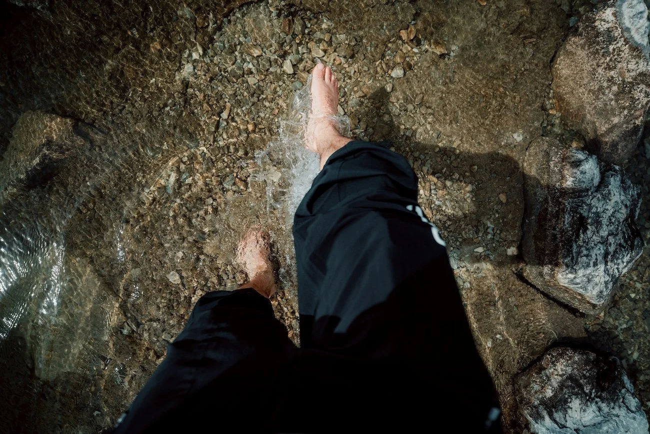 Person standing barefoot in clear, shallow water over a rocky riverbed.
