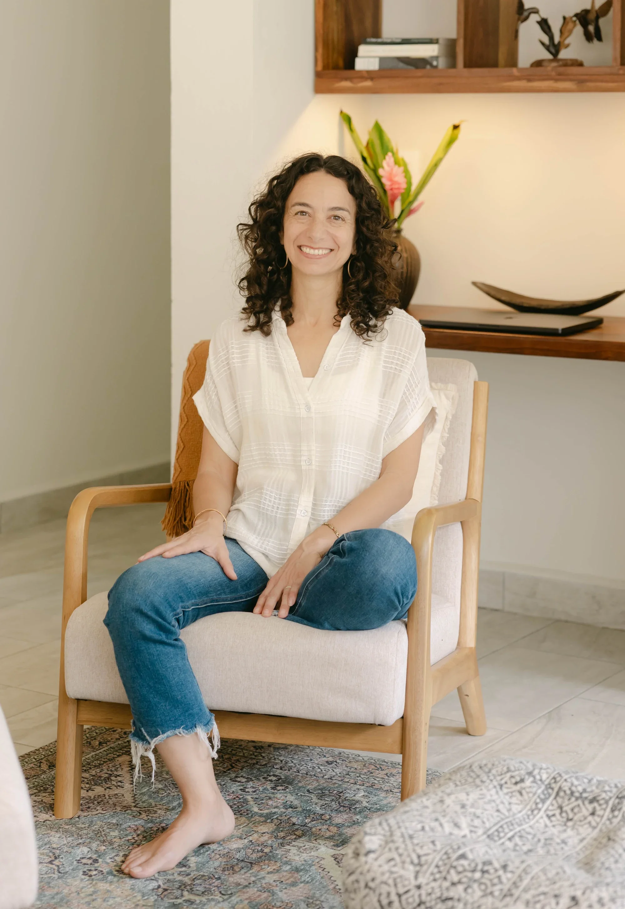 Alexis Lowenstein, psychotherapist, seated in her office. She is wearing a white blouse and blue jeans. Flowers, art and shelving are in the background.