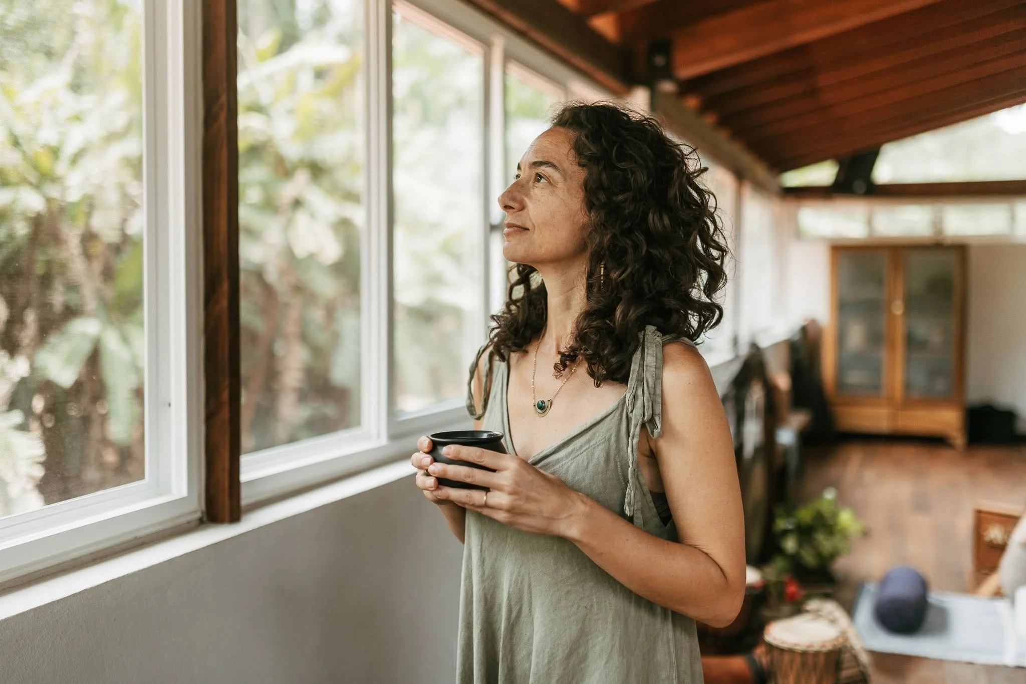 Alexis Lowenstein holding a black mug and looking out the window of a wooden interior room filled with natural light. She is at the Sattva Land Retreat Center in Belize.