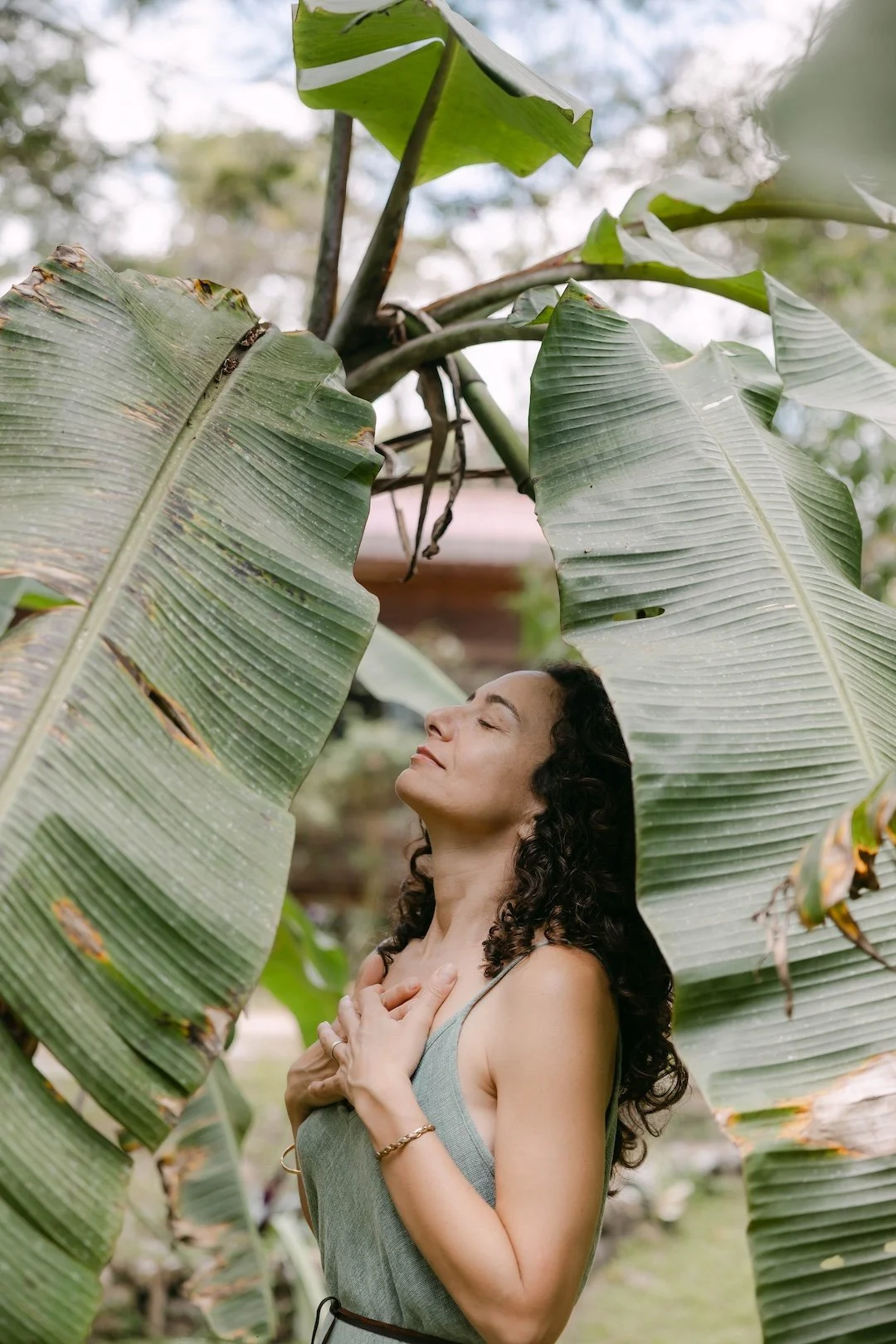 Alexis Lowenstein standing among large green leaves, with eyes closed and hands on her chest, appearing serene and content.