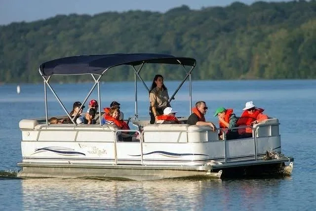 Group of people on a pontoon boat on a lake, with some wearing life jackets, surrounded by water and trees in the background.