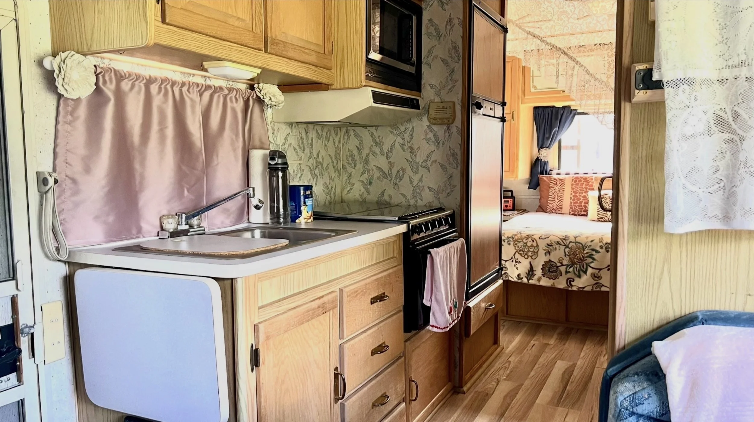 Interior of a vintage camper kitchen with wooden cabinets, a small sink, stove, microwave, and a view into the sleeping area with a bed and curtains.
