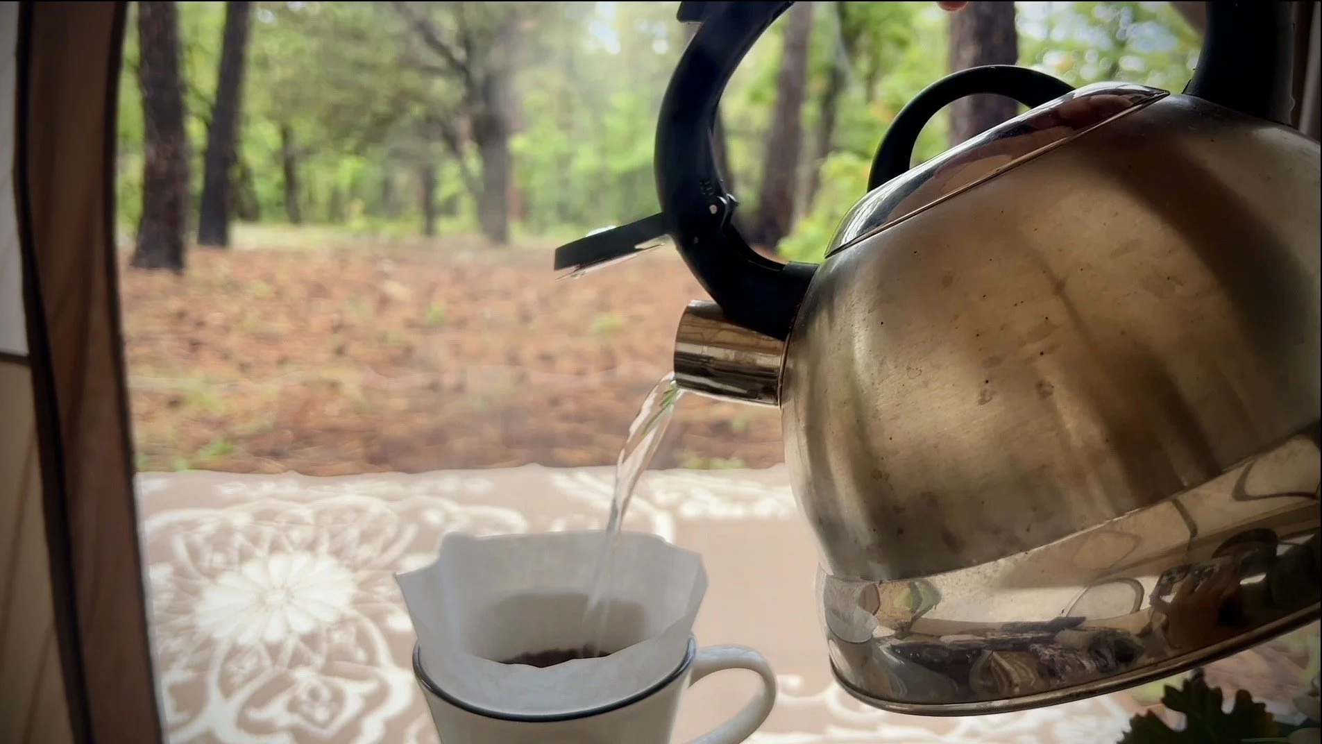 A kettle pouring hot water into a coffee filter placed in a mug, with a forest scene visible through a window in the background.