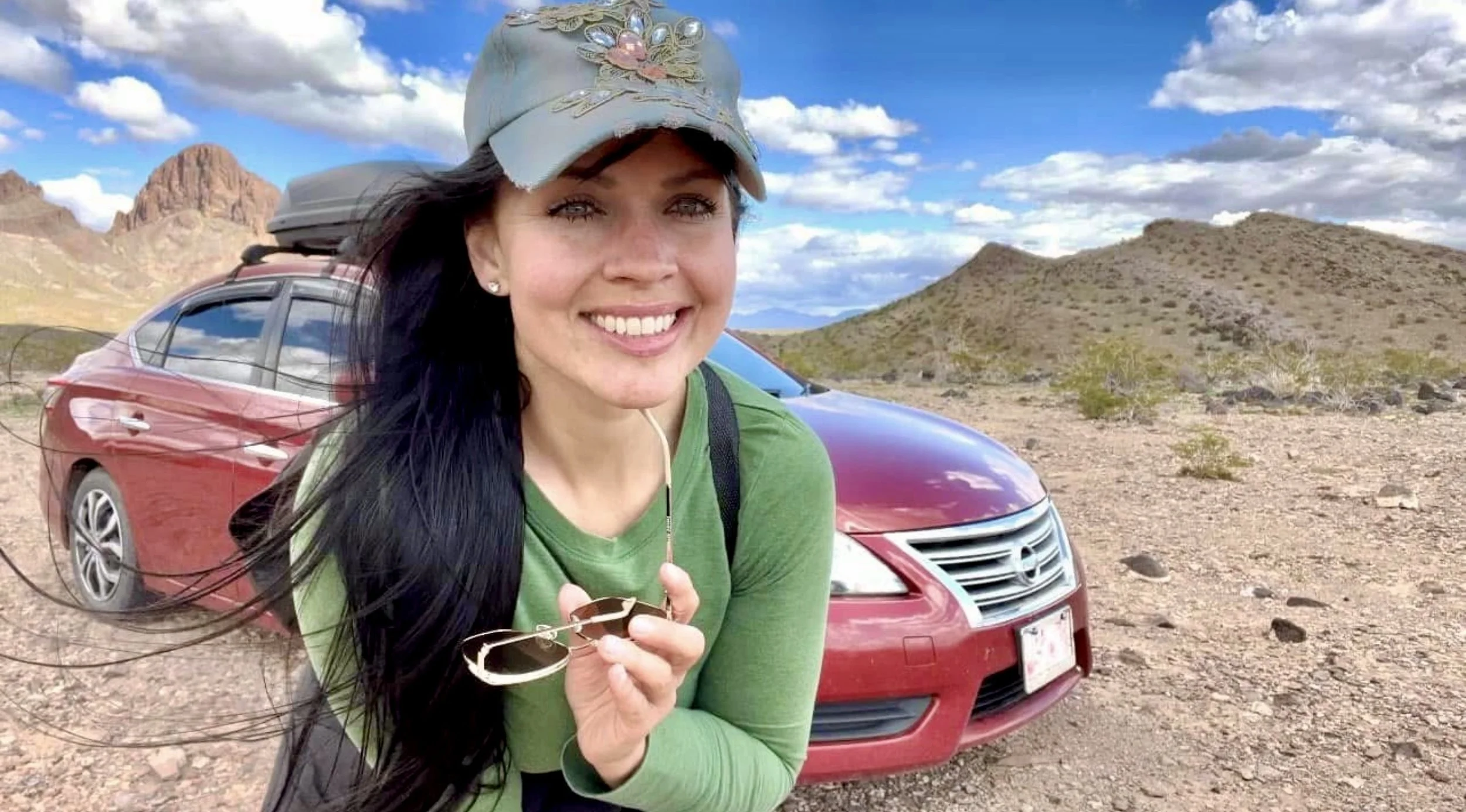 A smiling woman with long dark hair wearing a green shirt and a baseball cap, holding sunglasses, standing in front of a red car and a desert landscape with mountains and a partly cloudy sky.
