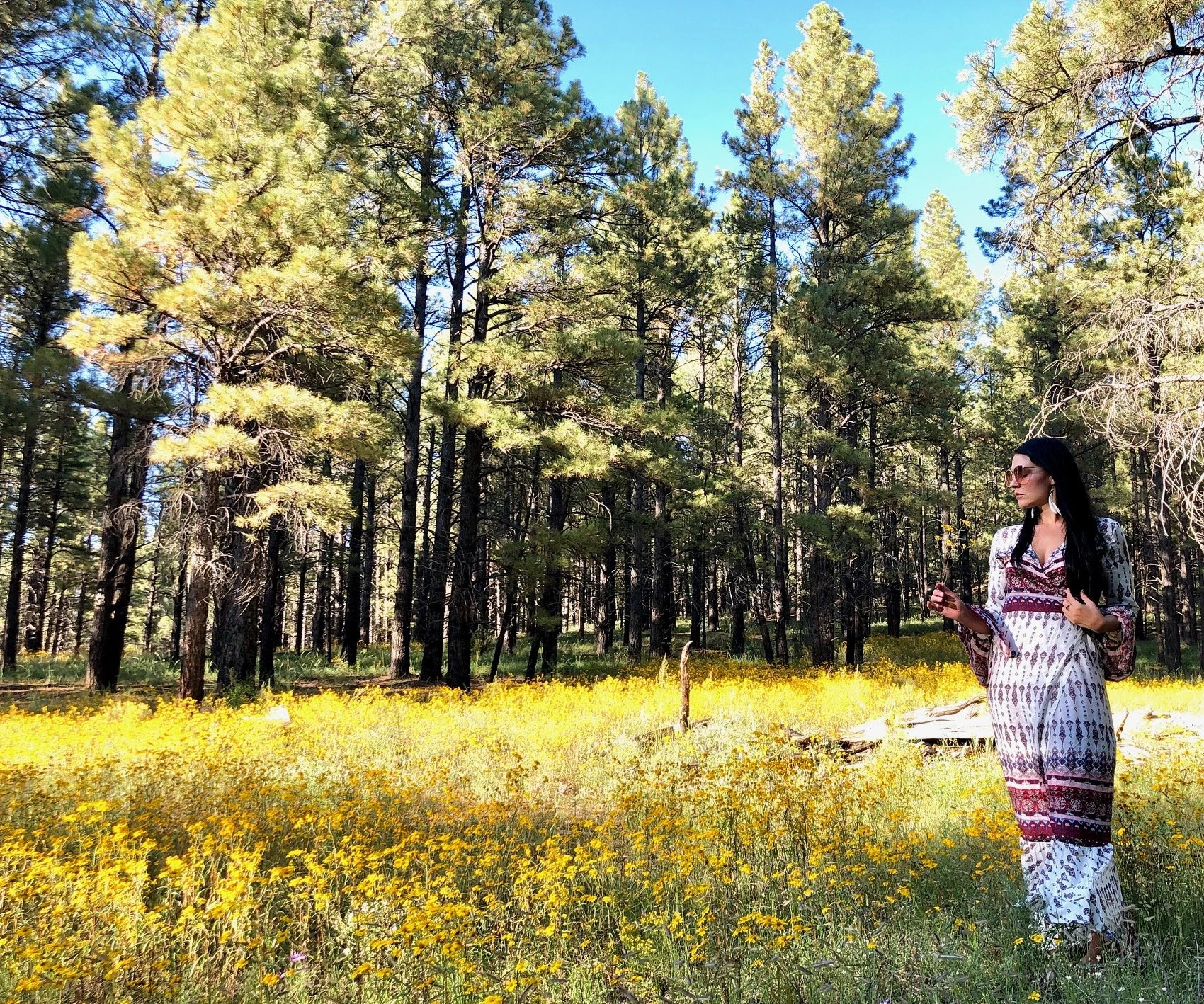 A woman in a patterned dress with sunglasses standing in a field of yellow flowers in a forest with tall trees and a blue sky.