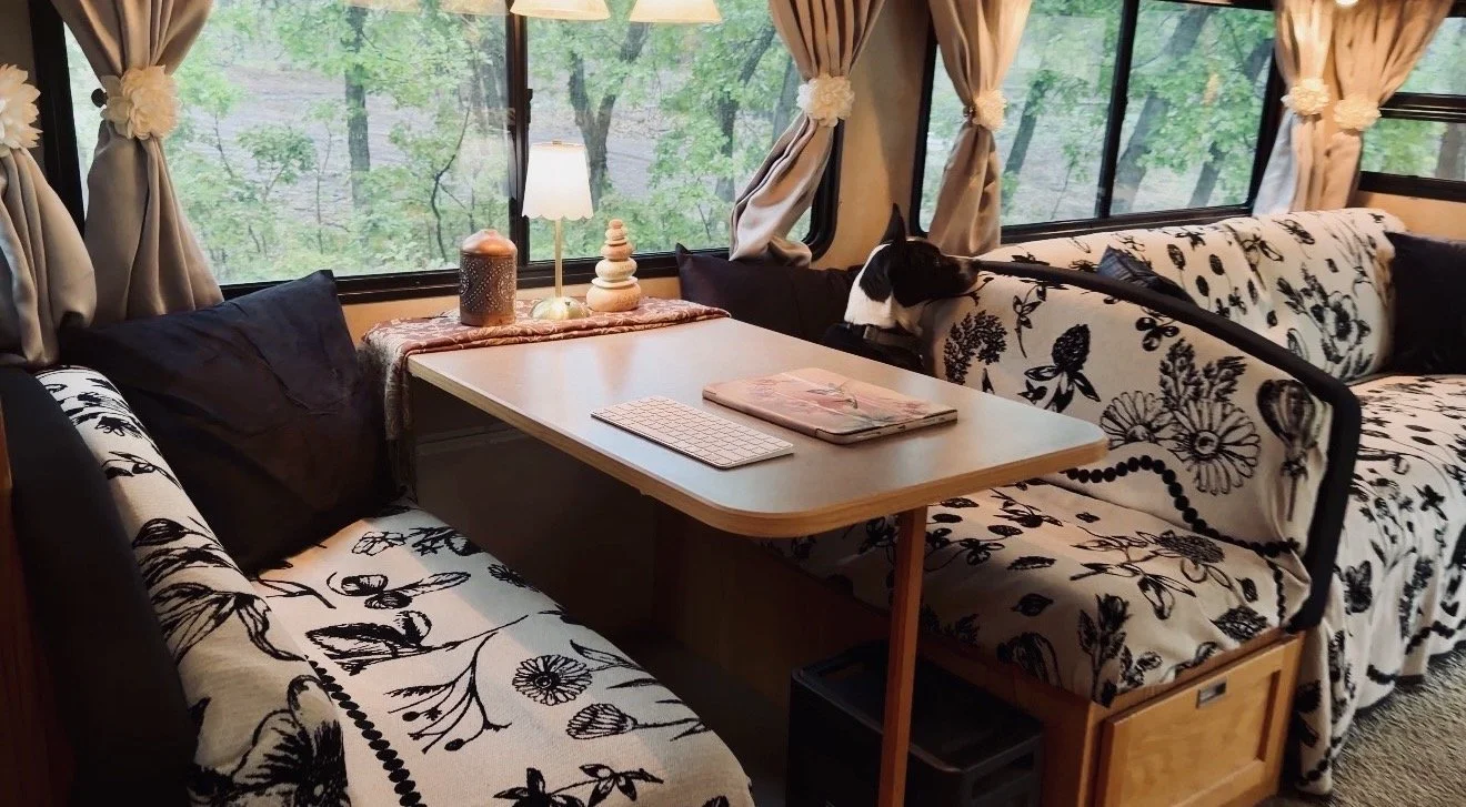 Interior of a cozy RV or camper with black and white floral-patterned cushions, a wooden table with a keyboard and magazines, and a dog sitting on a patterned cushion, looking out the window at green trees.