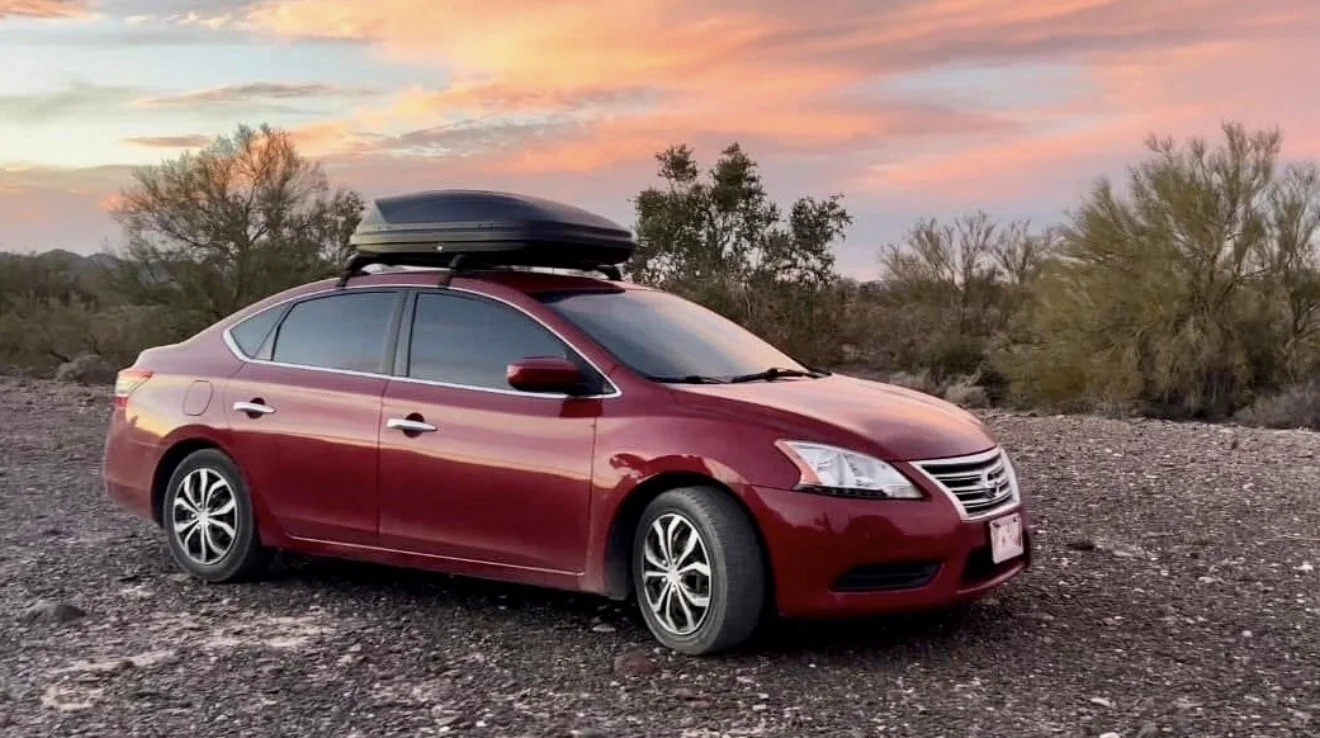 Red sedan car with a black roof cargo box parked on a gravel surface in a desert landscape at sunset, with sparse bushes and a colorful sky.