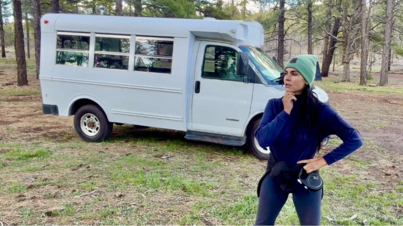 A woman stands outdoors in a forested area, wearing a green beanie and navy jacket, with a pensive expression, holding a camera in her waist. Behind her is a white van parked on grass.