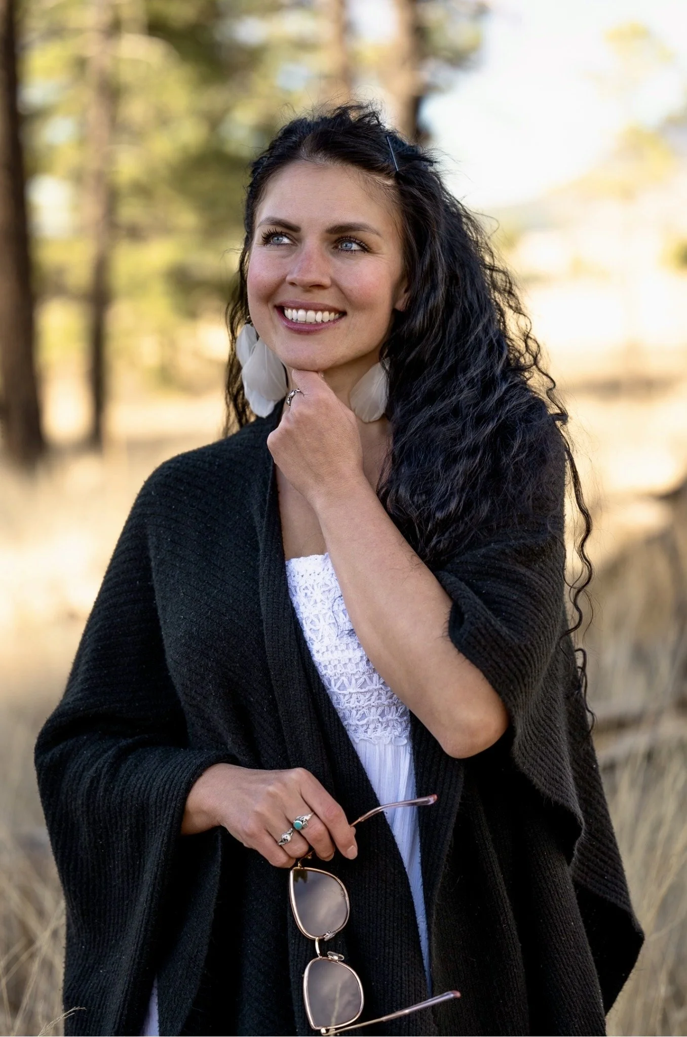 A woman with curly dark hair and blue eyes wearing a black sweater, white lace top, and holding sunglasses, standing outdoors in a field with trees in the background.
