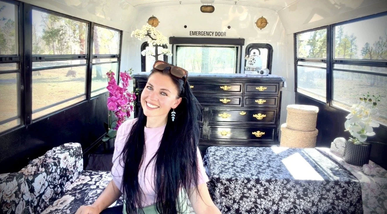 A woman with long black hair, sunglasses on her head, and earrings, smiling inside a decorated bus with large windows, floral decorations, a black dresser, and sunlight streaming in.