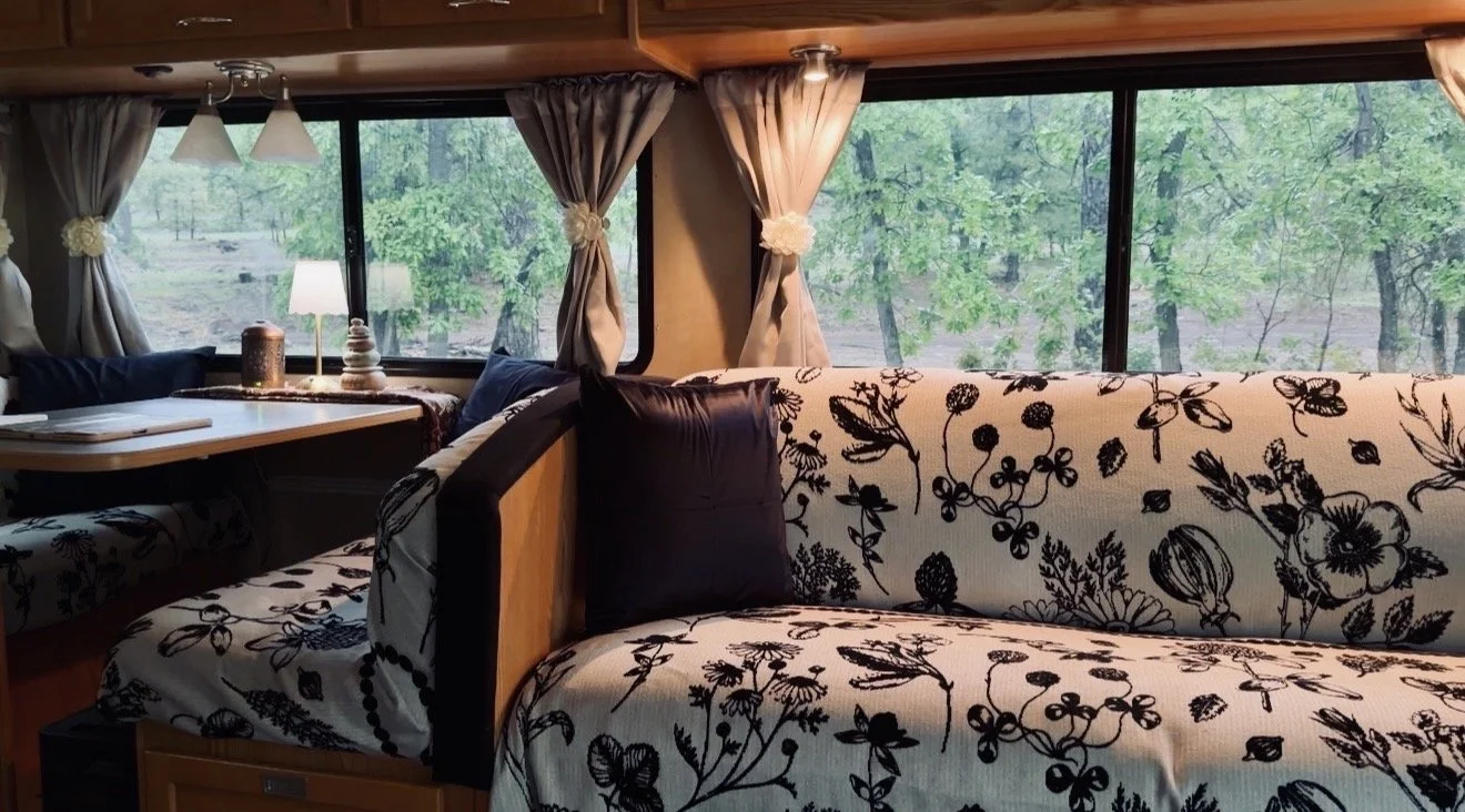 Interior of an RV with a floral-patterned sofa, large windows showing green trees outside, and a small dining area with curtains, a lamp, and a candle.