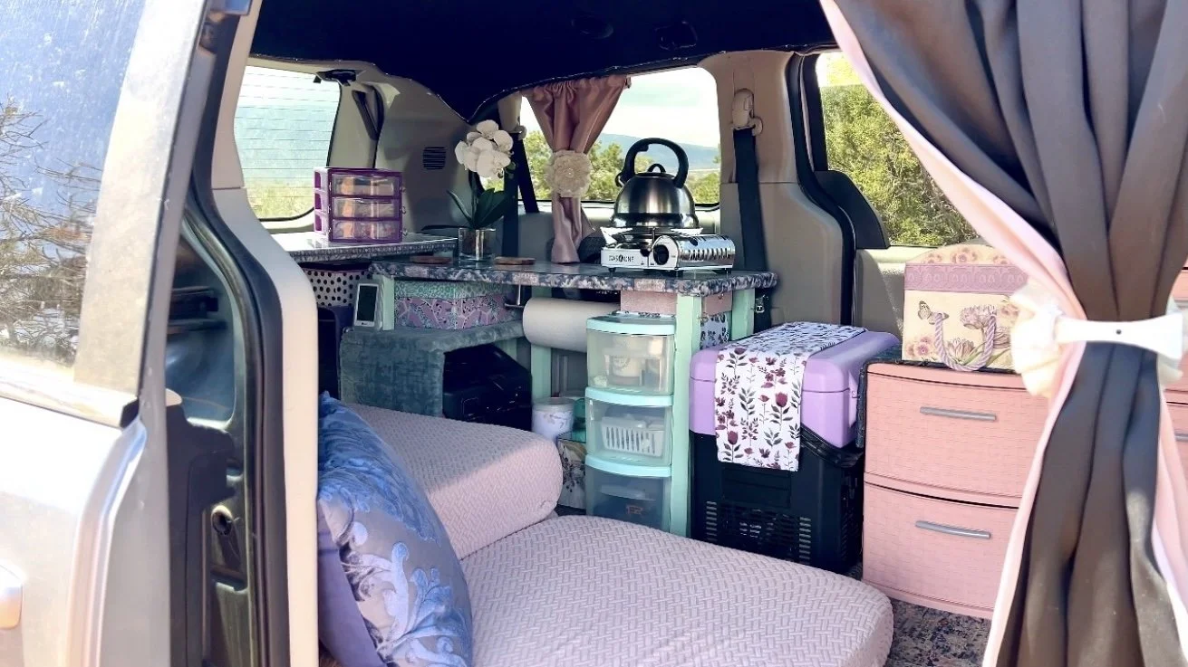 Interior of a converted van with a couch, storage drawers, a small kitchen area with a stainless steel kettle on a portable stove, and decorative curtains and flowers, set against a backdrop of greenery outside the van.