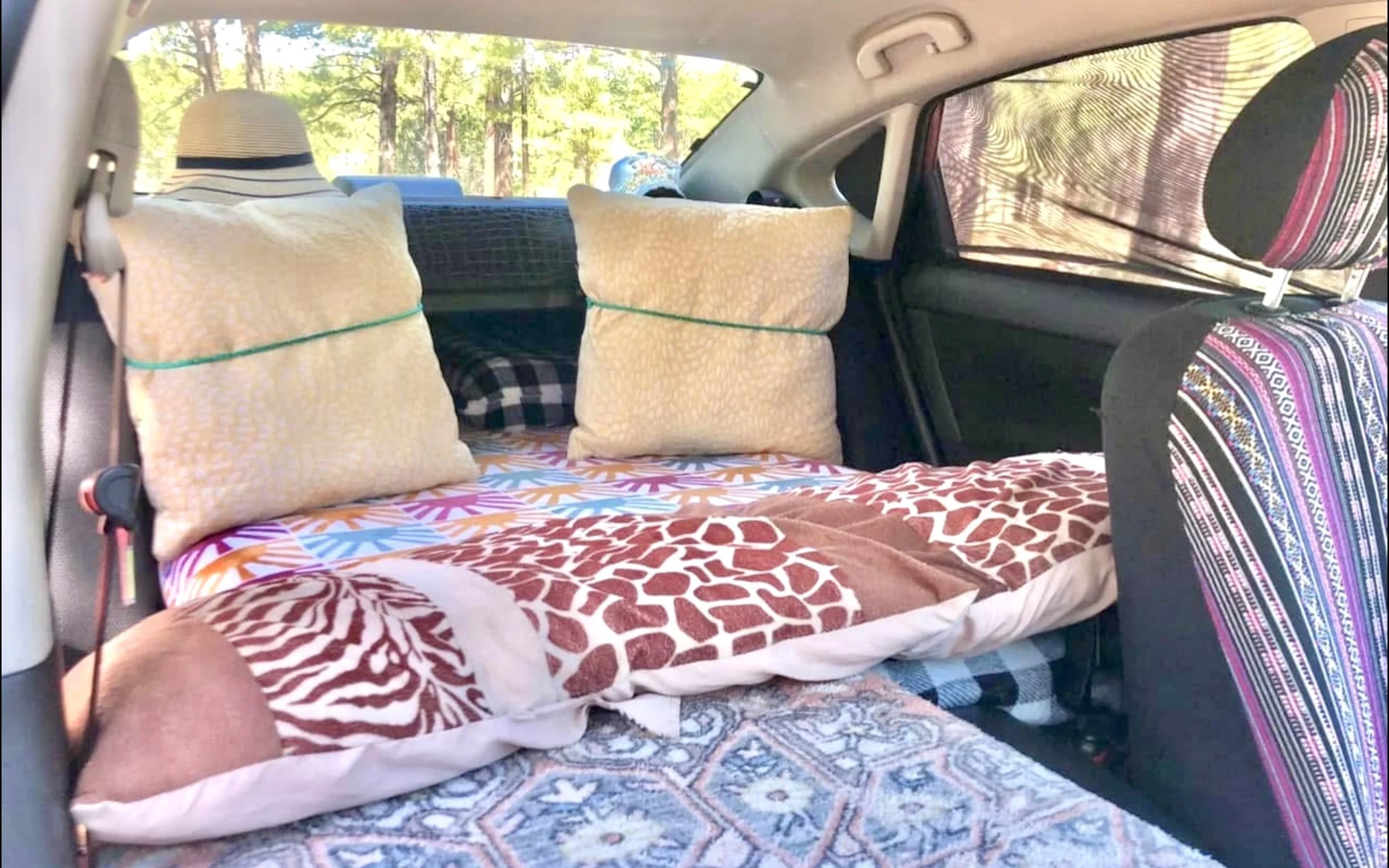 Interior of a vehicle with cushions and linens, including pillows with yellow and black patterns, a bedspread with a multicolored pattern, and curtains with intricate designs, set against a background of trees visible through the windows.