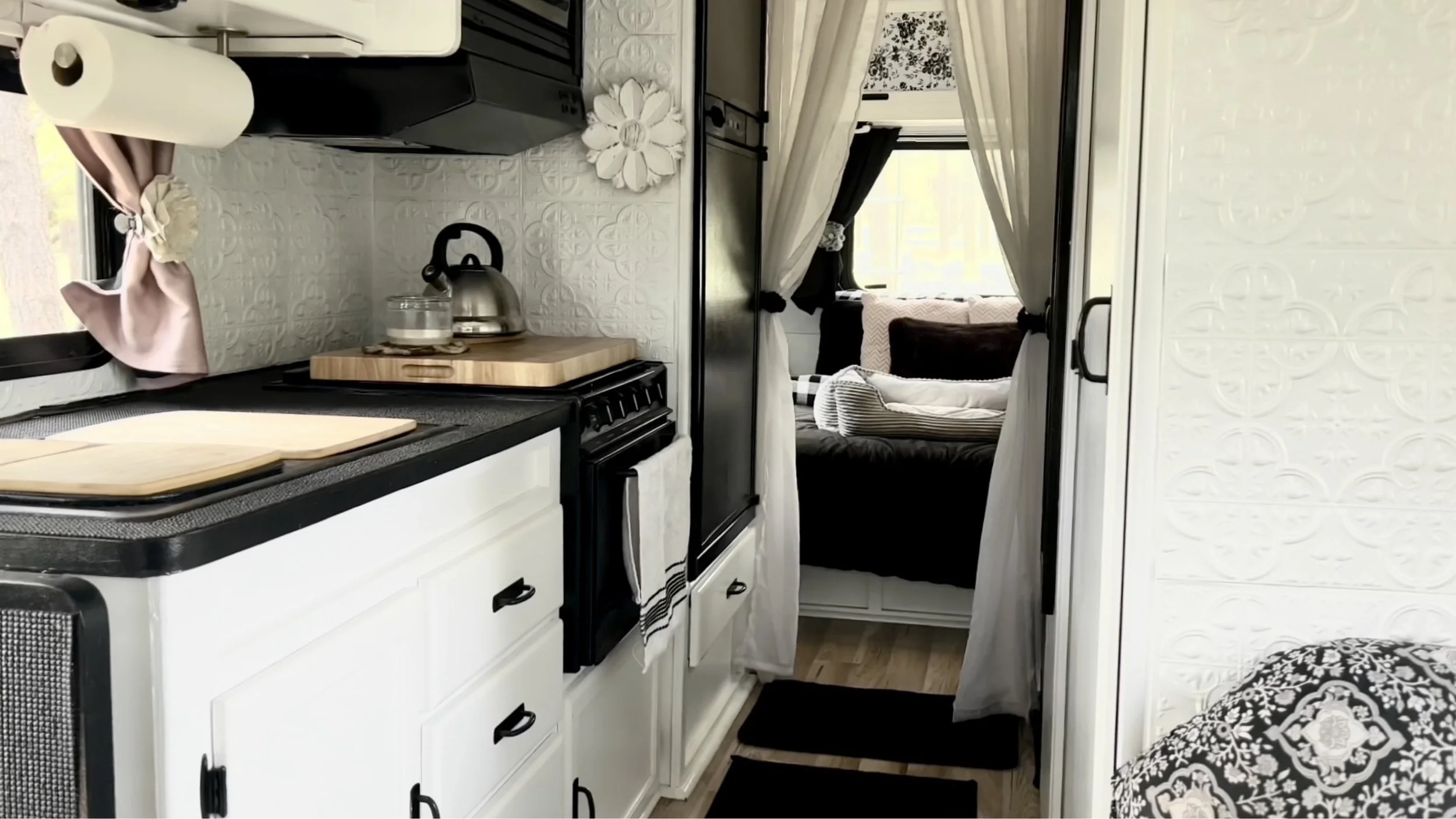 Interior view of a small kitchen with white cabinets, a stove, a kettle, and a window with curtains, leading to a living area with a sofa and other furniture.
