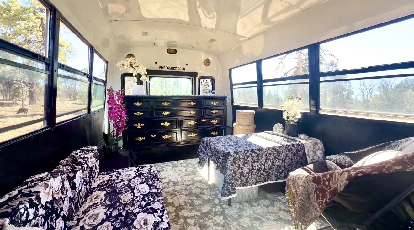 Inside a decorated bus with floral seating, a dresser, and flowering plants, windows showing trees outside.