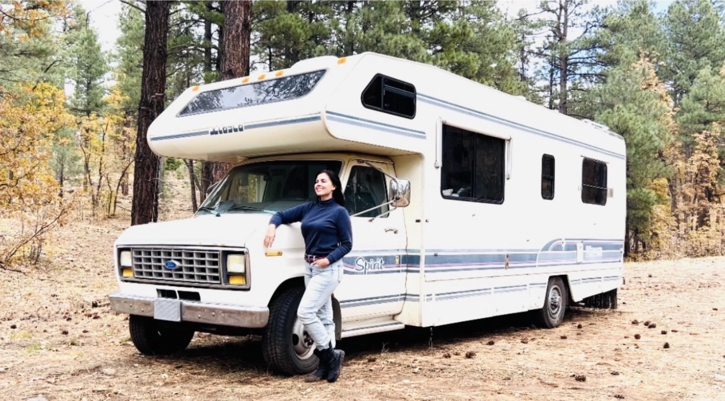 A woman in a navy blue top and light gray pants smiling with her arms crossed, standing beside a white vintage RV in a forested area with trees and yellow autumn leaves.