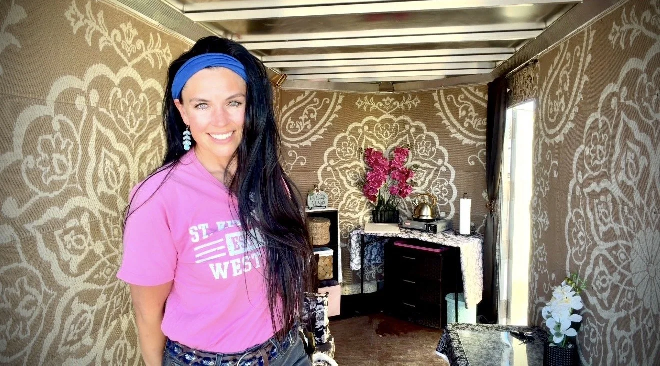 Smiling woman with long dark hair, wearing a pink T-shirt and a blue headband inside a decorated room with patterned wallpaper, flowers, and furniture.