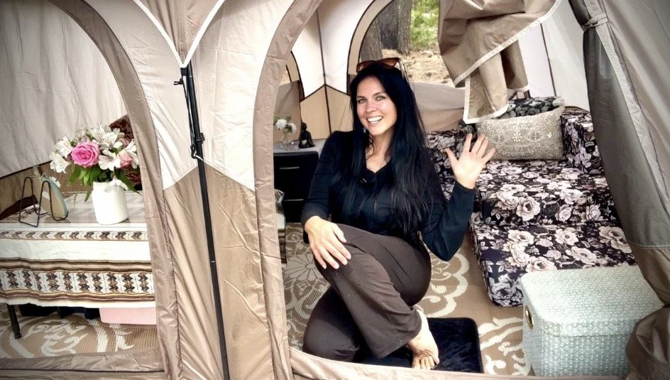 Woman smiling and waving inside a camping tent, with a bed, floral blanket, and a side table with a flower vase nearby.