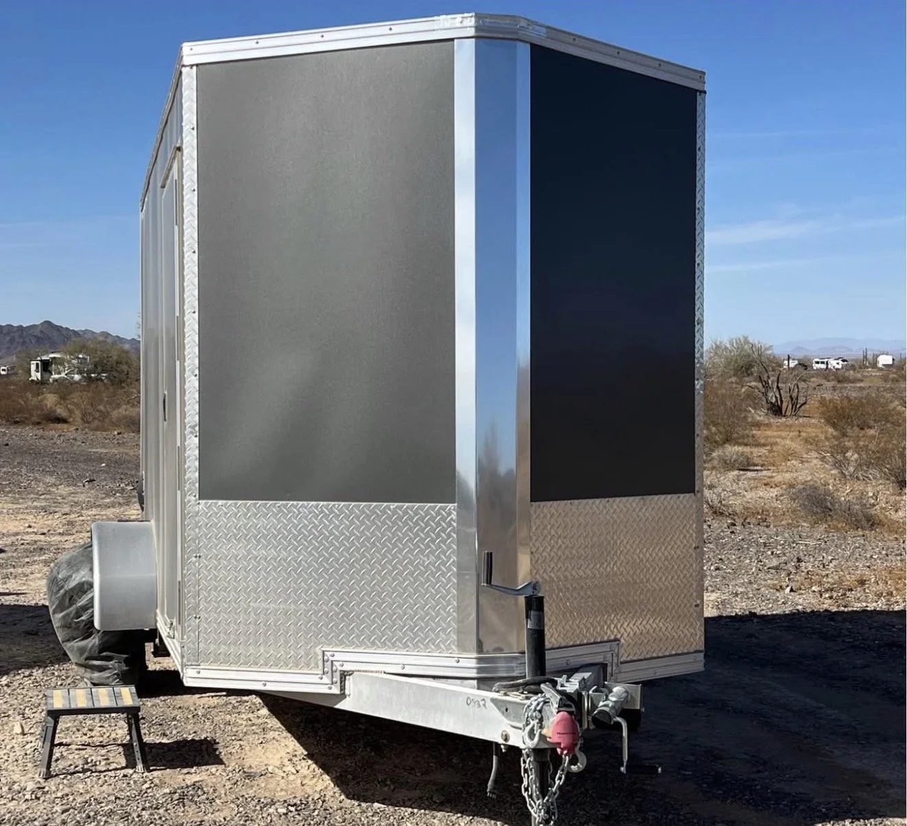 A silver, enclosed travel trailer with diamond plate trim along the bottom, parked on a desert landscape with mountains in the background. It has a small step stool beside it and an outdoor plug attached at the front.