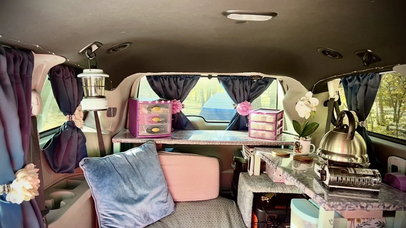 Interior of a decorated van with purple curtains, pink storage boxes, a silver kettle, a stove, and various decorative items like flowers and cushions.