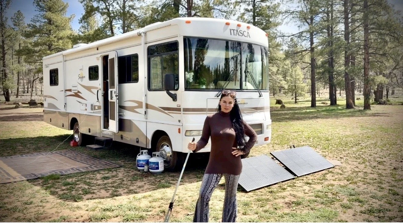 A woman standing in front of a white RV in a wooded area, holding a walking stick, with solar panels and propane tanks nearby.