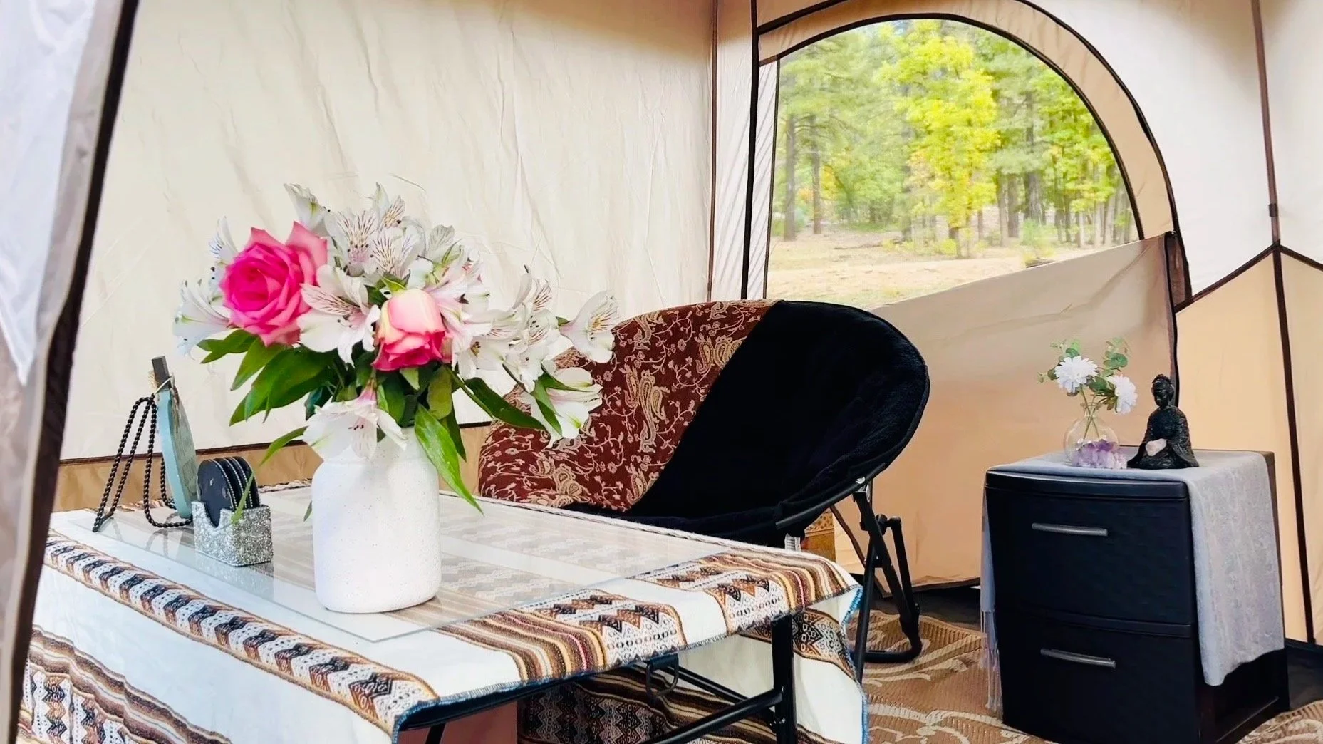 Inside a camping tent with a table decorated with a pink and white floral arrangement, a black chair with a patterned pillow, and a side table with a small Buddha statue, white flowers in a glass jar, and a purple crystal. Trees are visible outside through the tent window.