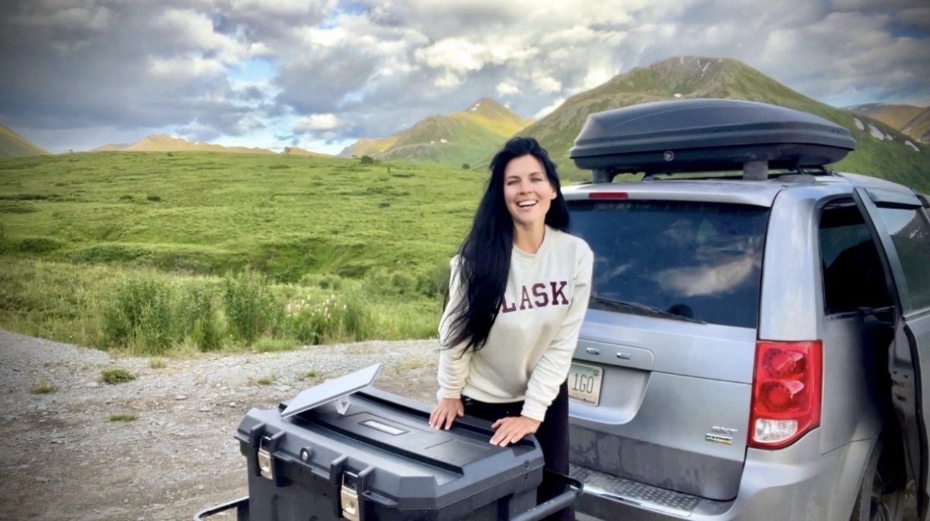 Woman smiling outdoors next to a silver SUV with a roof cargo box, in a scenic green mountainous landscape during daytime.