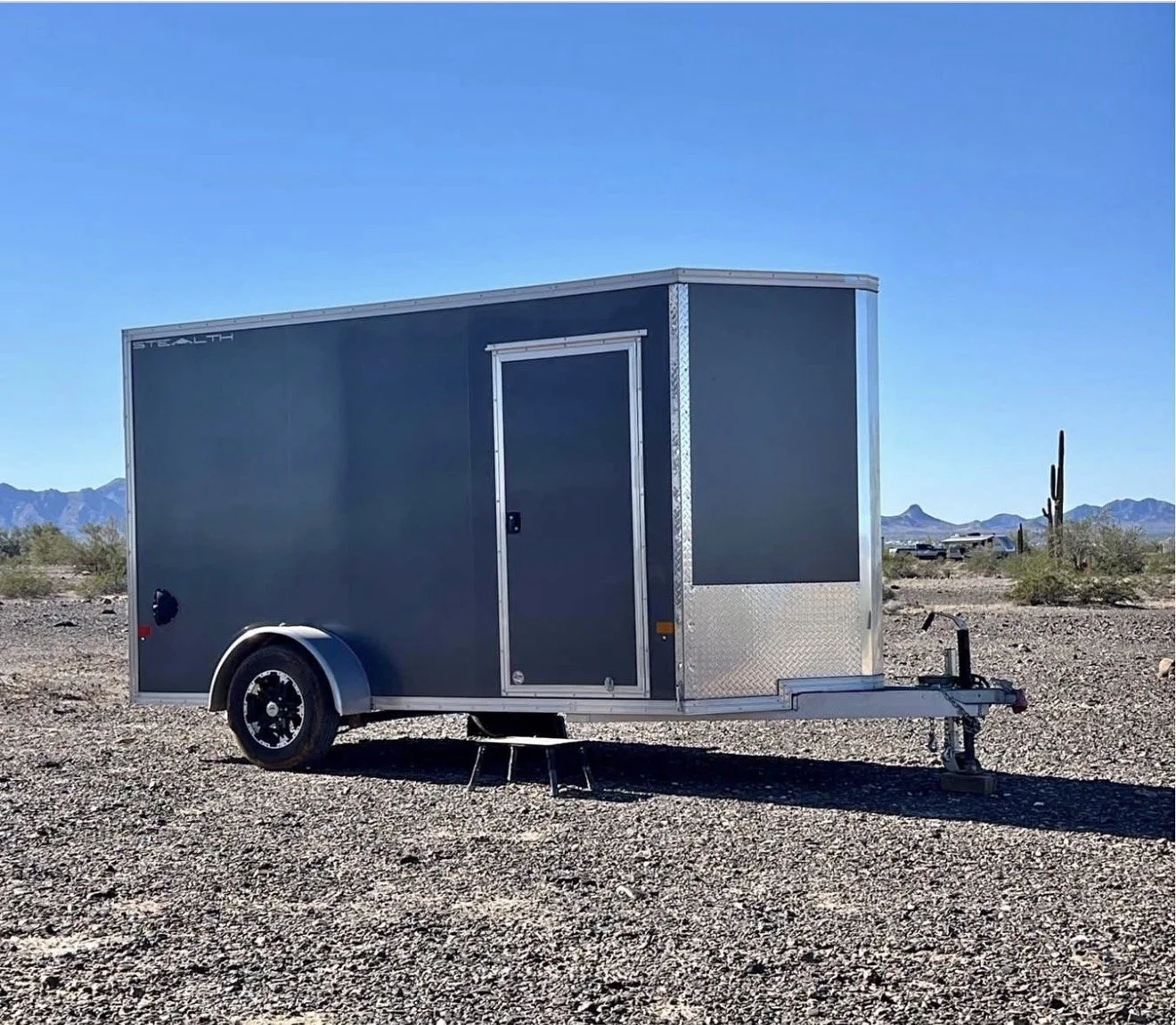 A dark gray enclosed utility trailer with a single wheel and a locked side door, parked on a gravel lot in a desert landscape with cacti, mountains, and a clear blue sky in the background.