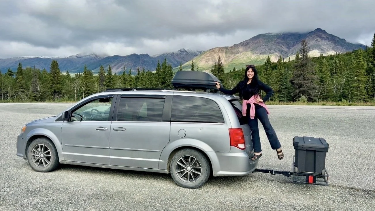 A woman with dark hair, wearing a black jacket, blue jeans, and sandals, is sitting on the back of a silver minivan, smiling. The minivan has a black cargo box on top and is attached to a trailer with a black storage container. The scene is set in a mountainous area with pine trees and a cloudy sky.