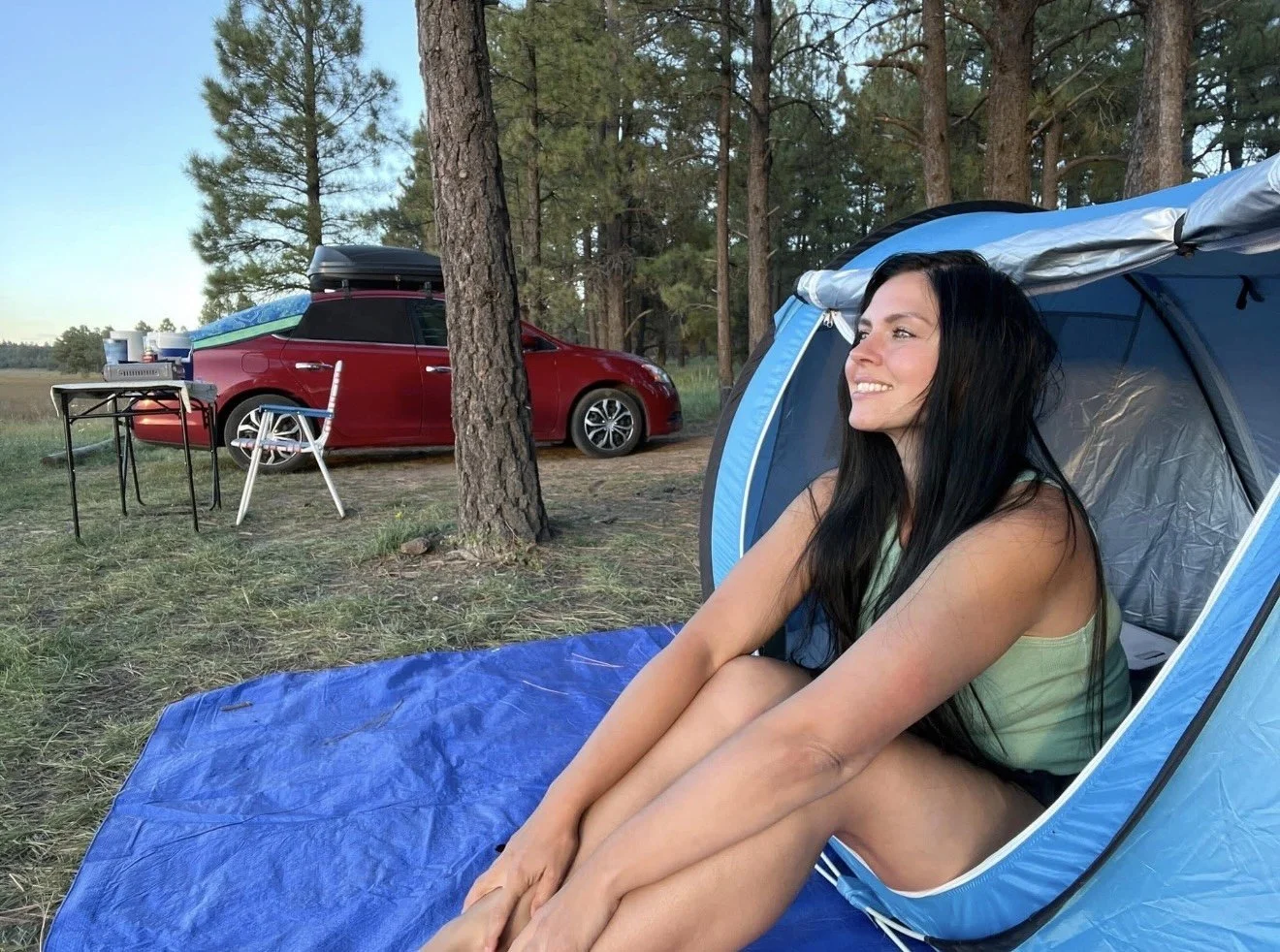 A woman with long black hair sitting inside a blue camping tent outdoors, smiling and looking to the right. In the background, there is a red car with a rooftop cargo box, a folding table, and a chair in a wooded area.