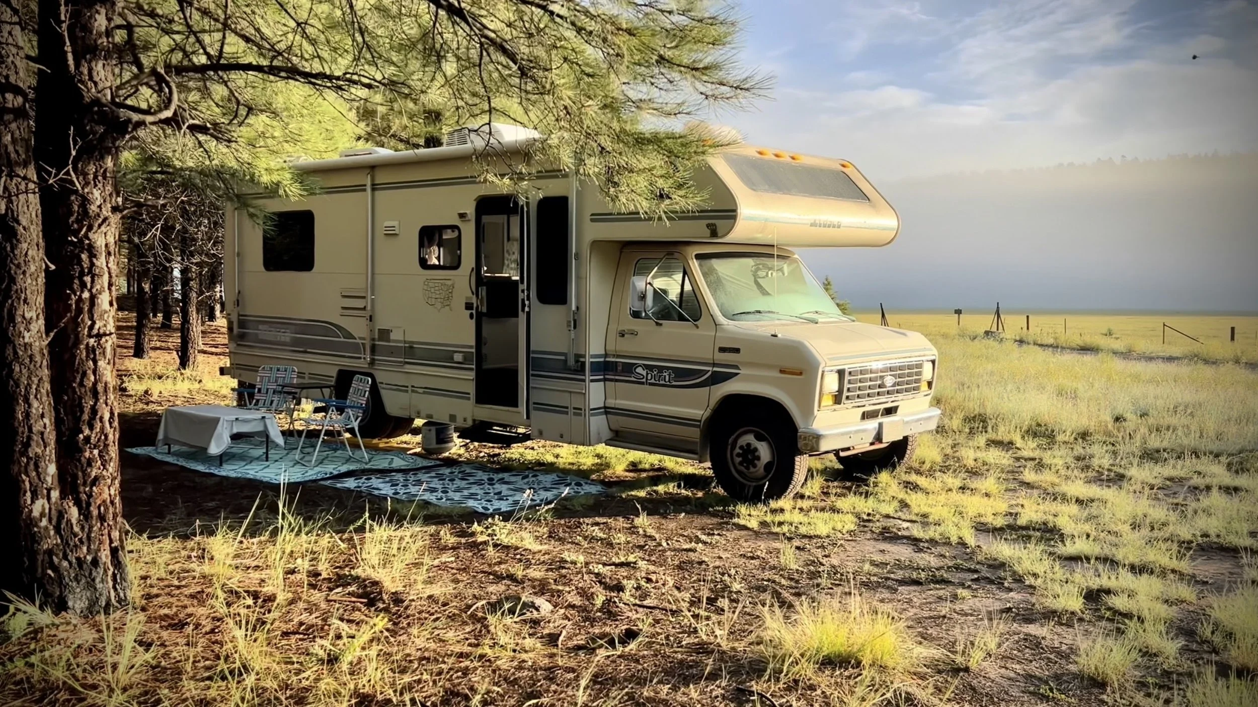 A beige RV parked in a grassy, wooded area with trees, a table, and chairs set up outside under a partly cloudy sky.