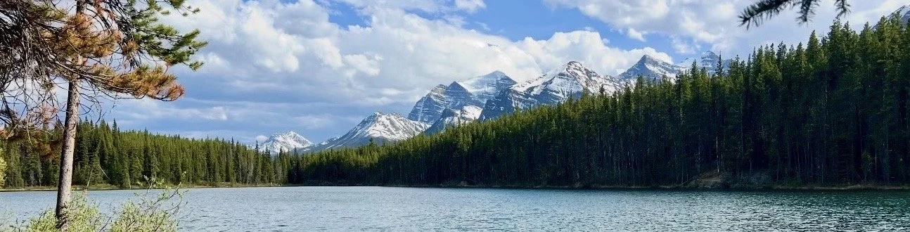 A serene lake surrounded by pine trees, with snow-capped mountains in the background under a partly cloudy sky.