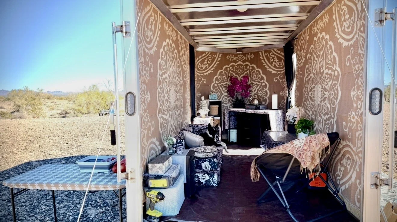 Interior of a decorated outdoor trailer or tiny home with patterned wallpaper, furniture, plants, and decorative items, in a desert landscape