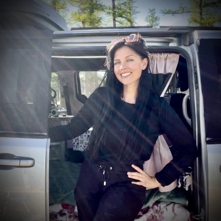 A woman smiling and posing outside a camper van in a forested area.