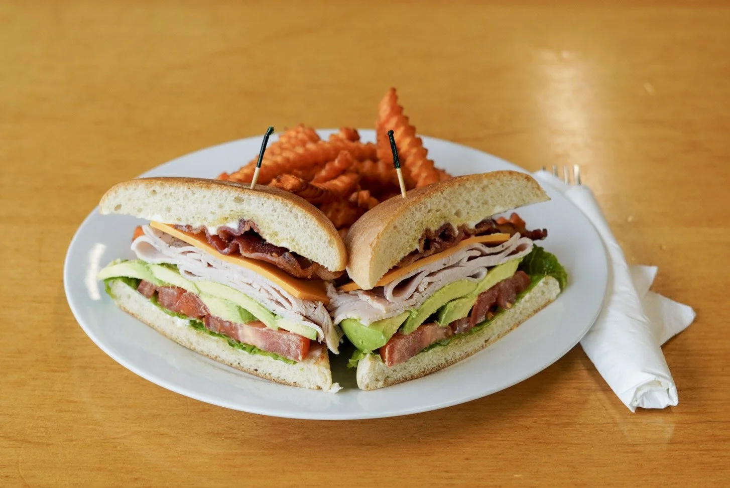 A plate with a submarine sandwich cut in half, filled with turkey, bacon, cheddar cheese, lettuce, and tomato, served alongside waffle fries.