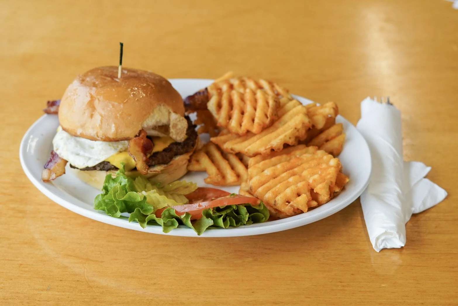 Plate with cheeseburger, waffle fries, and waffle cut sweet potato fries, with a side of lettuce, tomato, and a slice of onion, on a wooden table with a wrapped roll nearby.