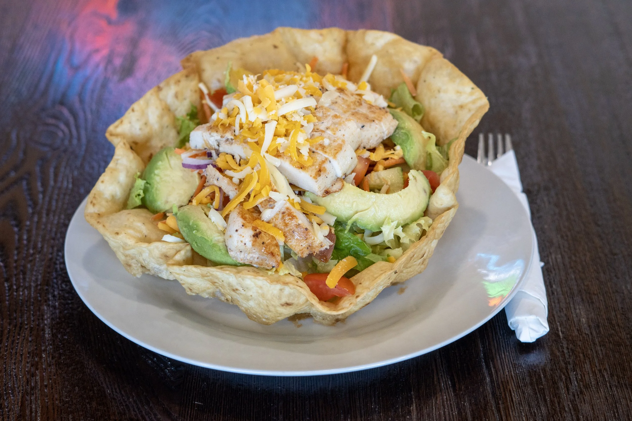 A taco salad served in a crispy taco bowl with grilled chicken, shredded cheese, lettuce, avocado slices, and diced tomatoes, placed on a white plate on a dark wooden table.