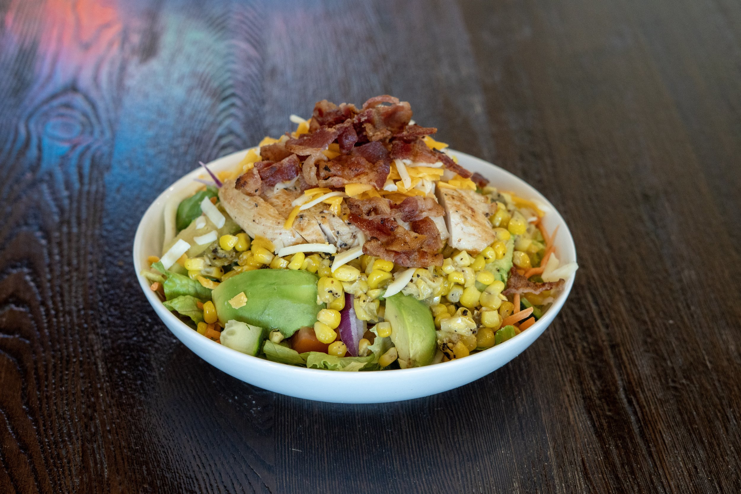 Bowl of salad topped with bacon pieces, shredded cheese, grilled chicken slices, and mixed vegetables including corn, carrots, and lettuce, on a dark wooden table.