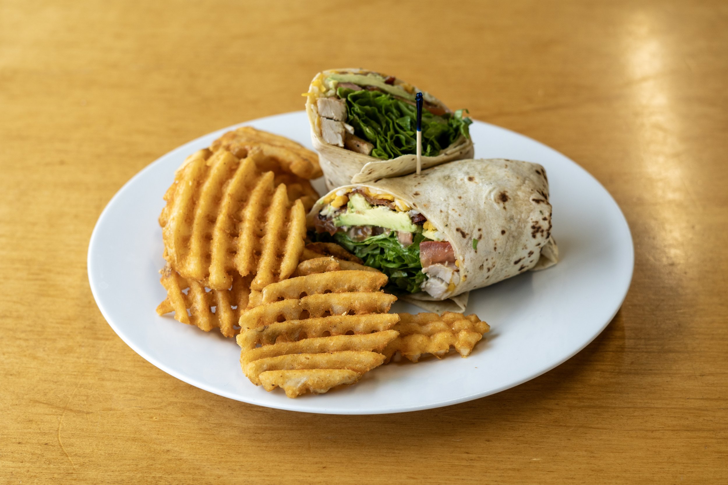 A white plate with waffle-cut French fries and a chicken wrap with lettuce, tomato, cheese, and grilled chicken, garnished with a toothpick, on a wooden table.