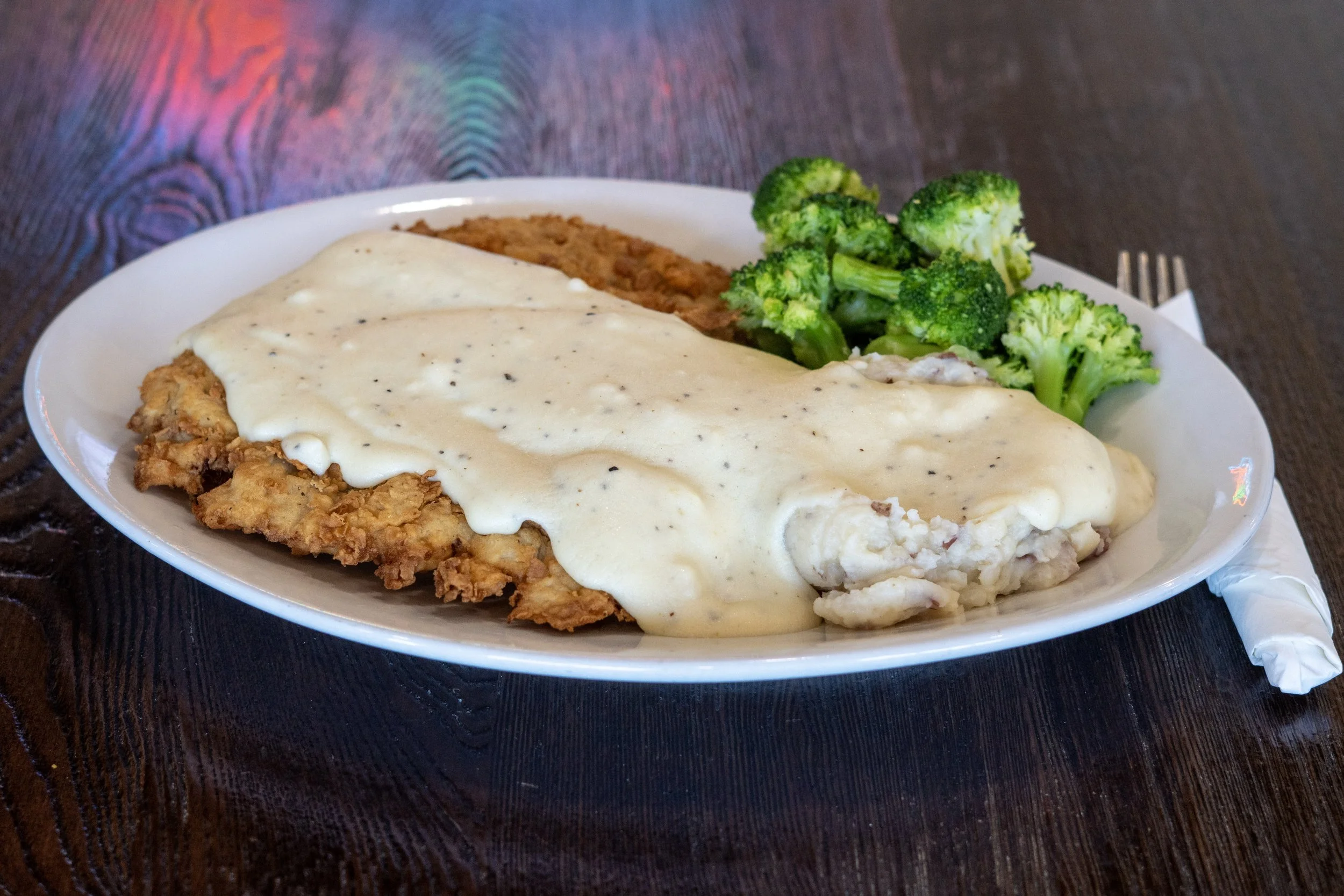 A plate of food with fried chicken topped with creamy white gravy, served with steamed broccoli and a serving of baked casserole on a dark wooden table, with a fork on the right side of the plate.