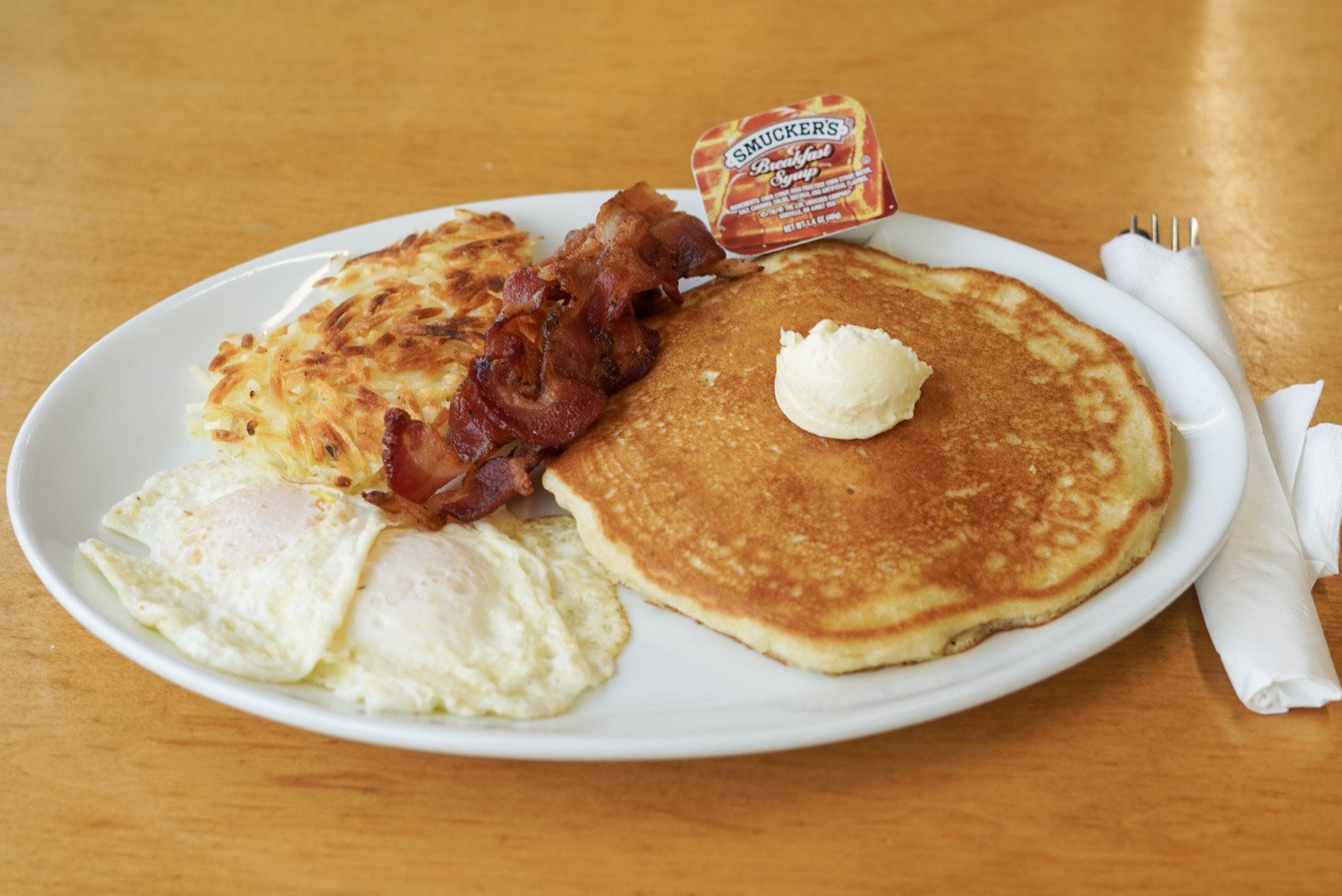A breakfast plate with eggs, hash browns, bacon, pancakes with butter, and a packet of syrup on a wooden table.