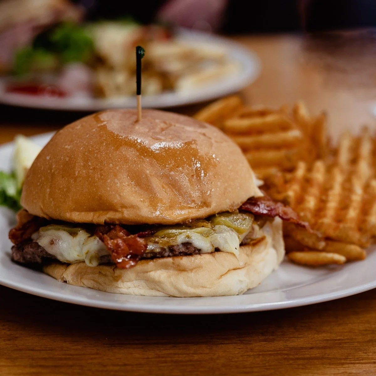 A cheeseburger with bacon, lettuce, and melted cheese in a bun, served with waffle fries on a white plate.