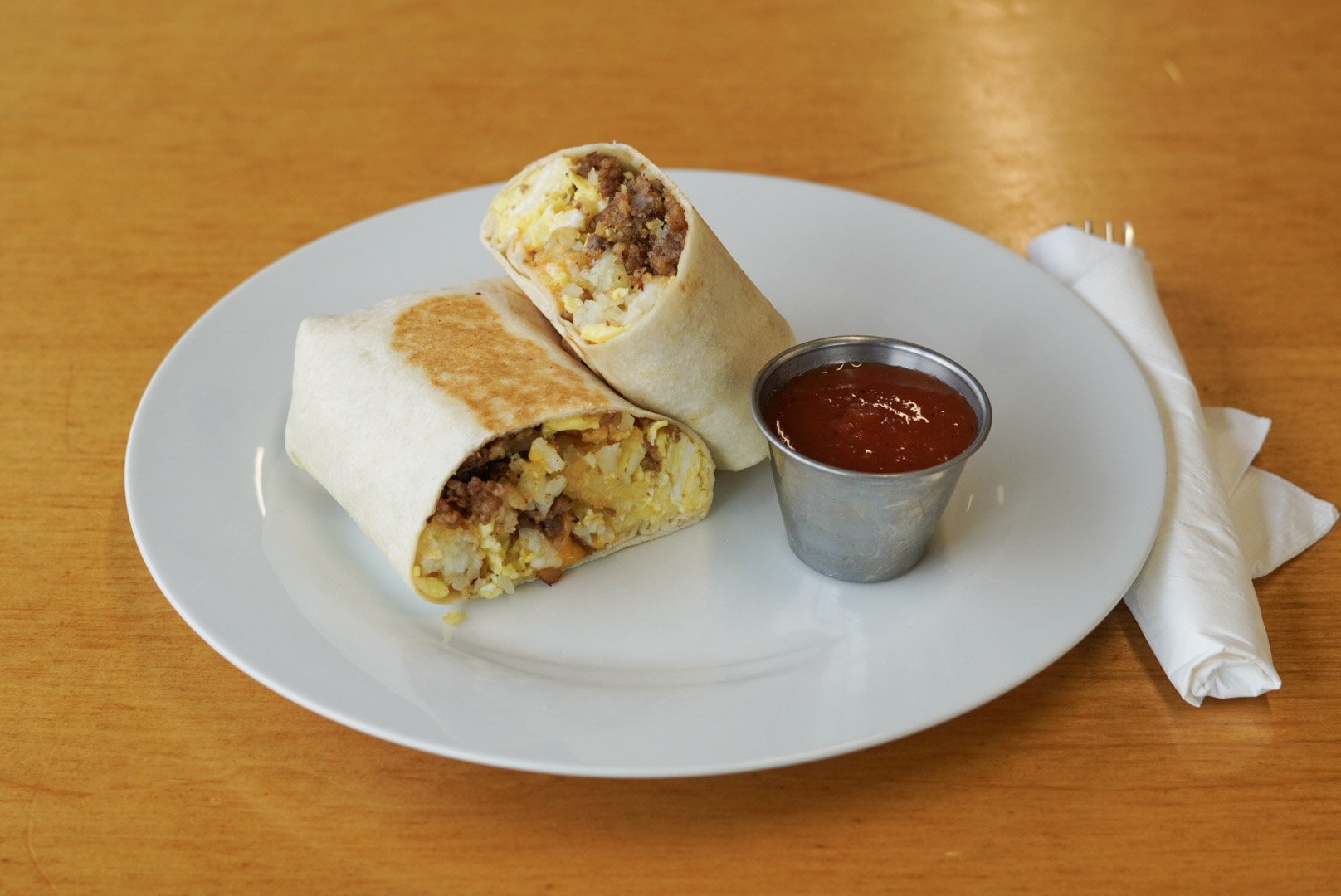 A white plate with two breakfast burritos cut in half, filled with scrambled eggs, sausage, and cheese, accompanied by a small cup of salsa, on a wooden table with a wrapped napkin and fork.