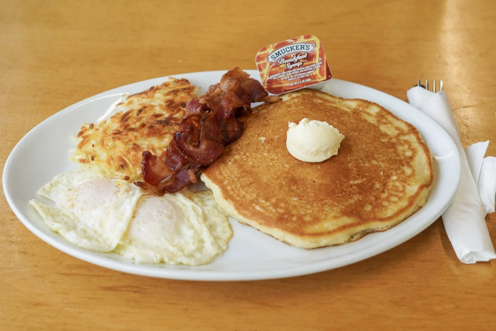 A breakfast plate with crispy hash browns, sunny side up eggs, bacon, and a stack of pancakes with butter, accompanied by butter and syrup on a small packet labeled 'Smucker's.' napkin with silverware wrapped inside is on the side.