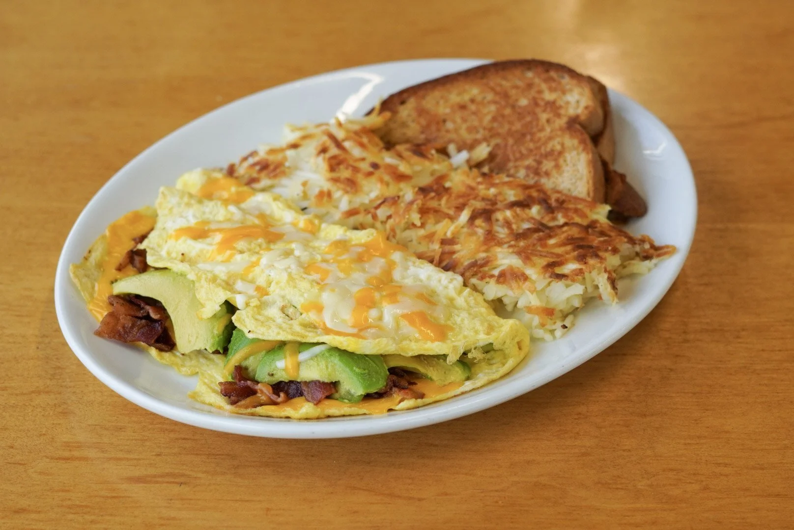 A white oval plate with scrambled eggs topped with melted cheese, avocado, and bacon, along with hash browns and two slices of toasted bread, on a wooden table.