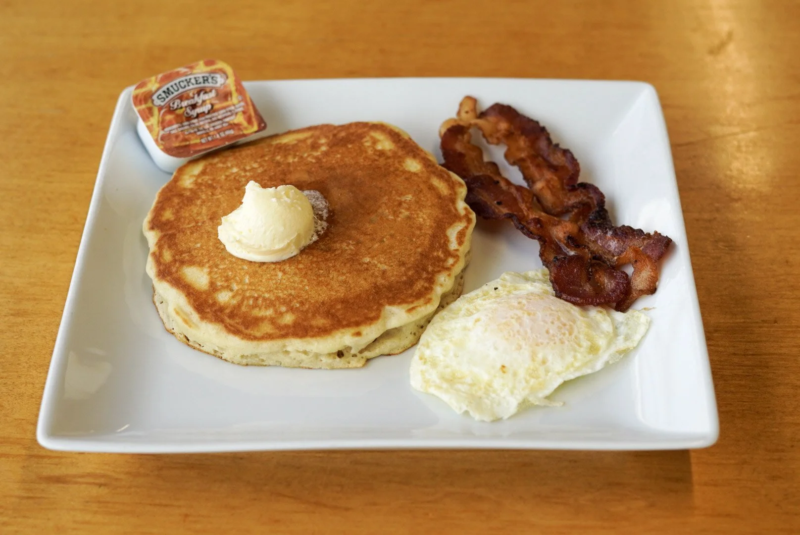 Plate of breakfast food including pancakes with butter, fried egg, bacon, a small container of syrup, and a piece of toast on a wooden table.