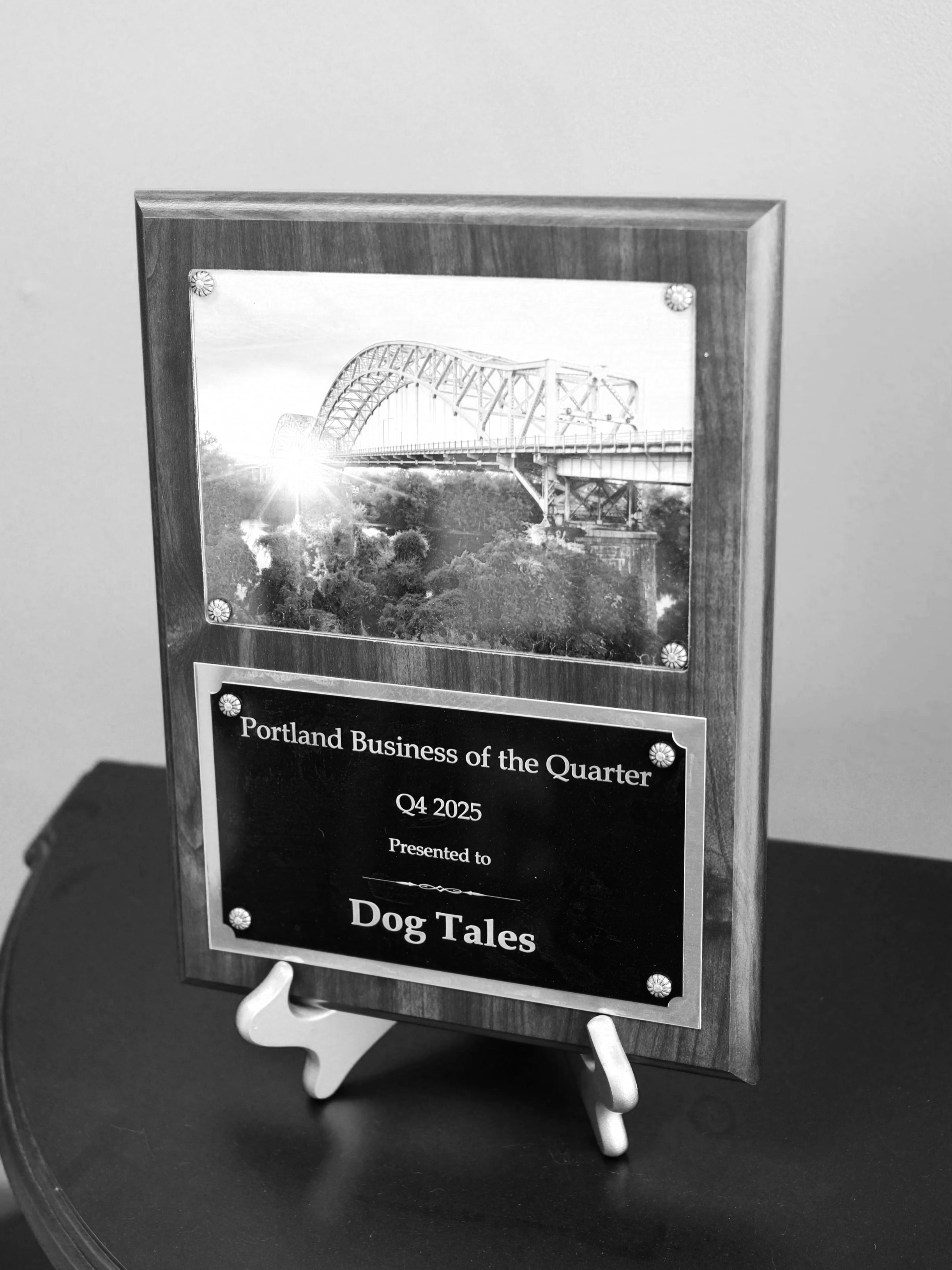 A framed photograph of a bridge, placed on a white stand, with a plaque indicating it is for Portland Business of the Quarter, Q4 2025, titled 'Dog Tales'.